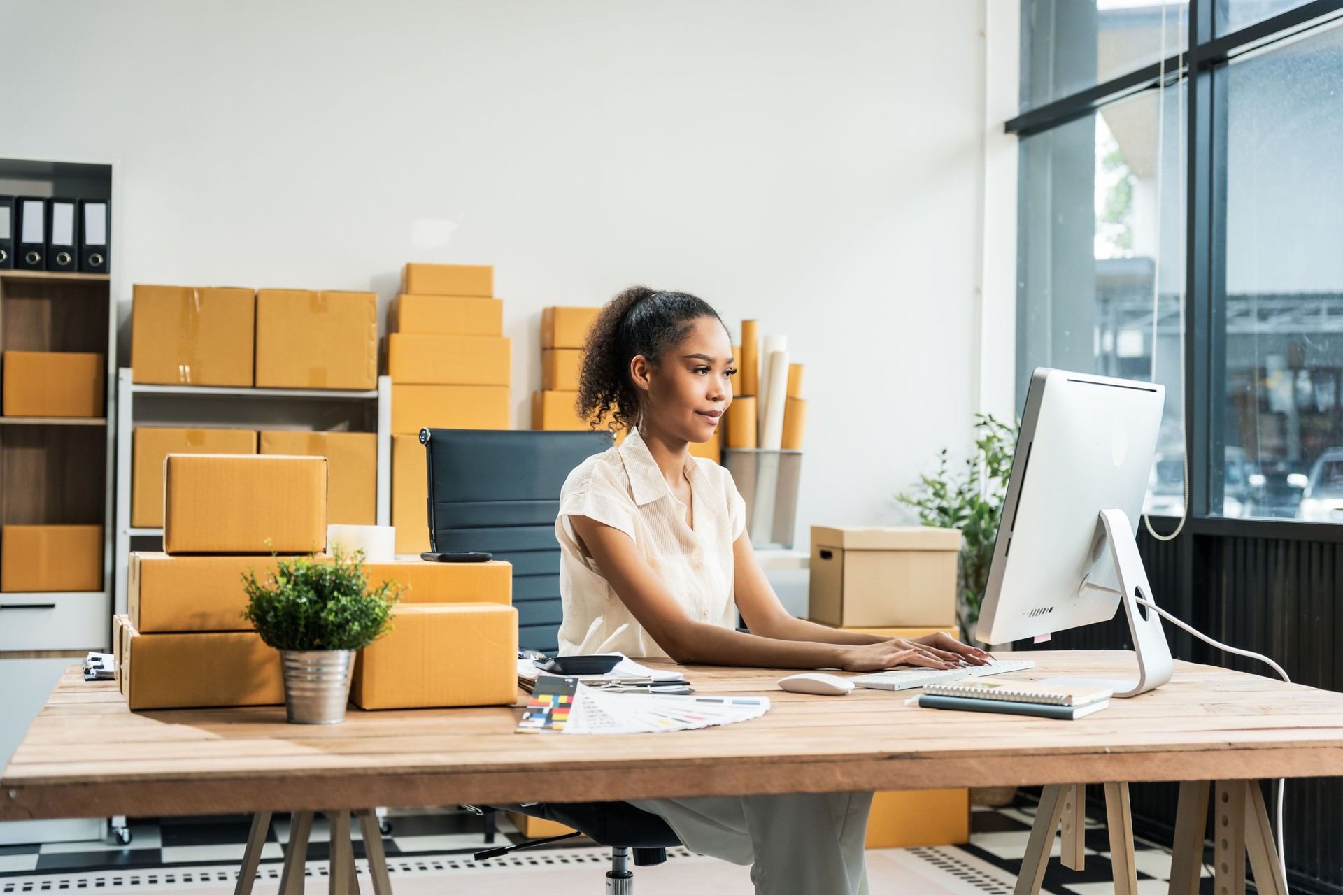 An African American woman with an afro works at a wooden desk, equipped with a laptop, desktop computer, and parcel cardboard boxes, focusing on SME logistics, inventory, and online orders.